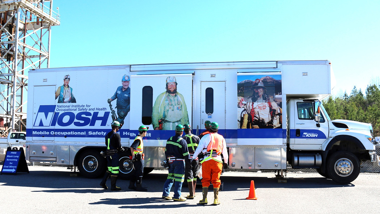 NIOSH's Mobile Occupational Safety and Health Unit parked outdoors, with several workers in safety gear standing in front of the vehicle. The truck displays large images of miners and the NIOSH logo. A sign nearby advertises free black lung screenings for coal miners.
