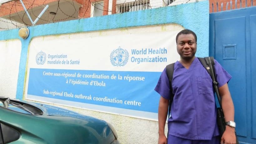Black man in scrubs standing in front of a World Health Organization poster