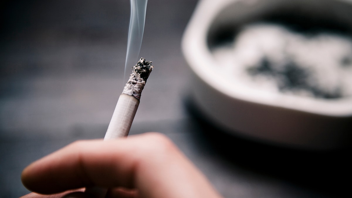 a hand holding a cigarette in the foreground, and a blurry ashtray in the background.