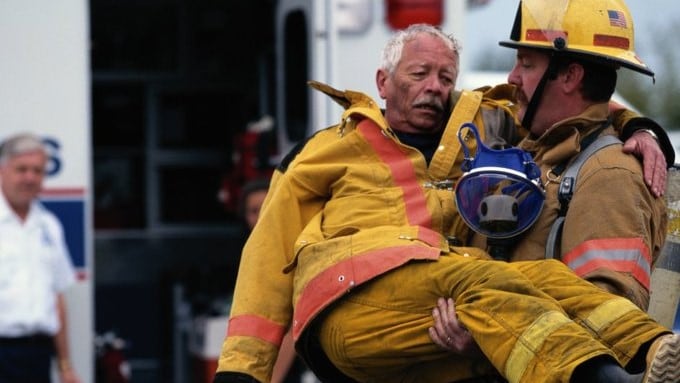A firefighter carries an older firefighter to an ambulance