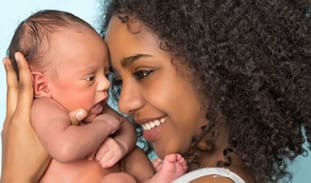 A smiling mother with curly hair lovingly holds her newborn baby, both facing each other closely.