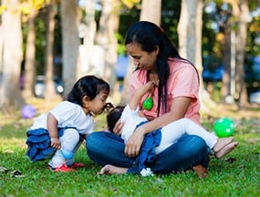 Mom breastfeeding her baby in a park while her toddler kisses the baby’s head.