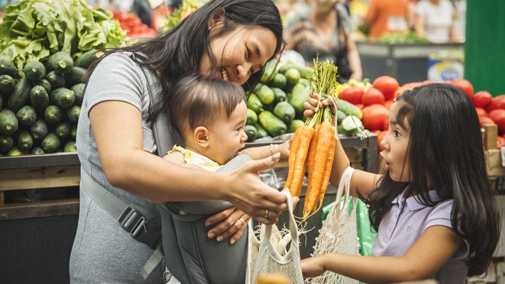 Mother with two young children at a produce store. One child is putting a bunch of carrots in a shopping bag.