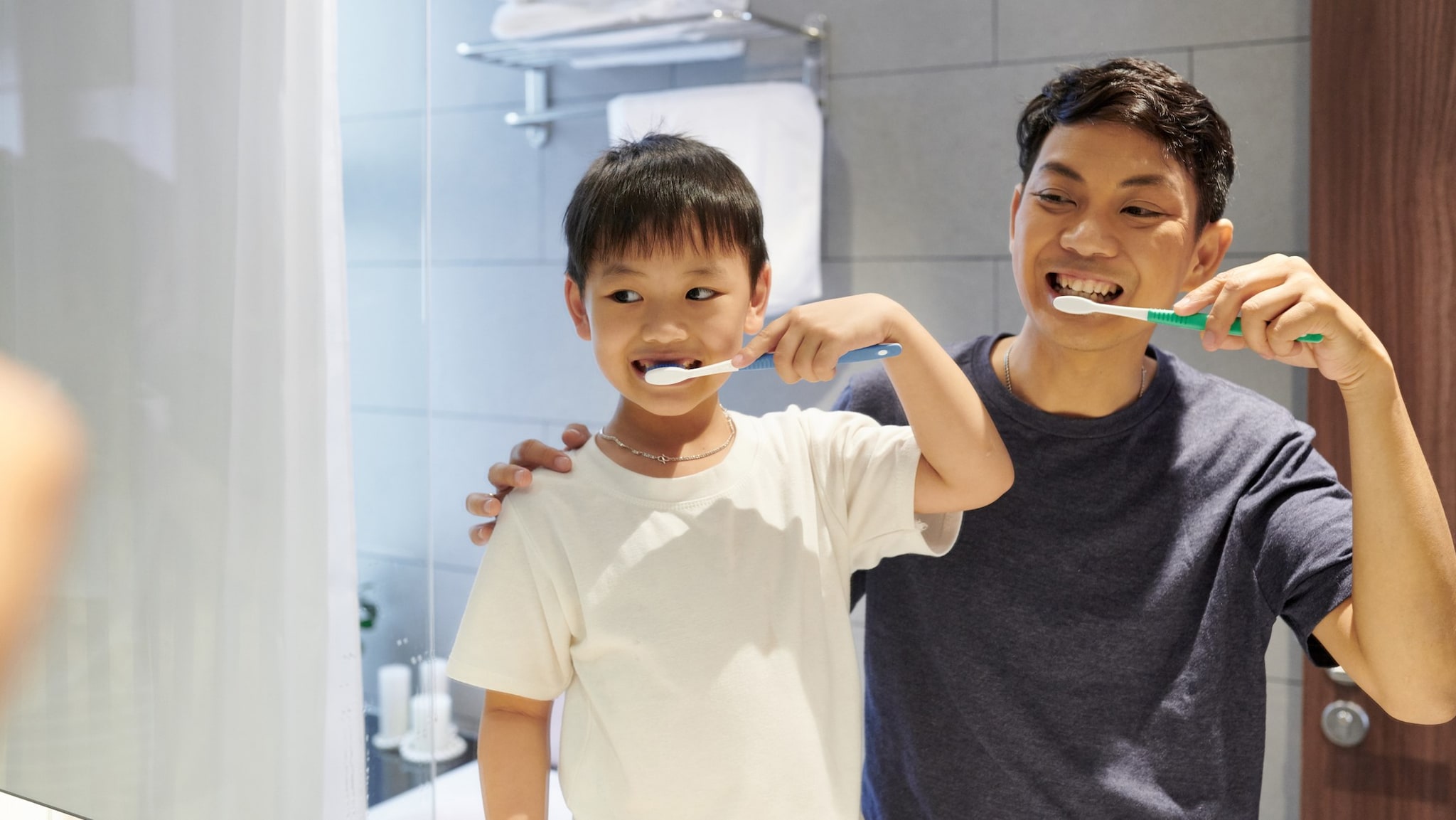 Father and son brush their teeth while looking in bathroom mirror.