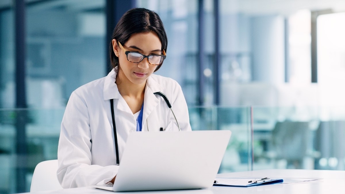 A healthcare provider wearing glasses sits at a table with an open notebook computer.