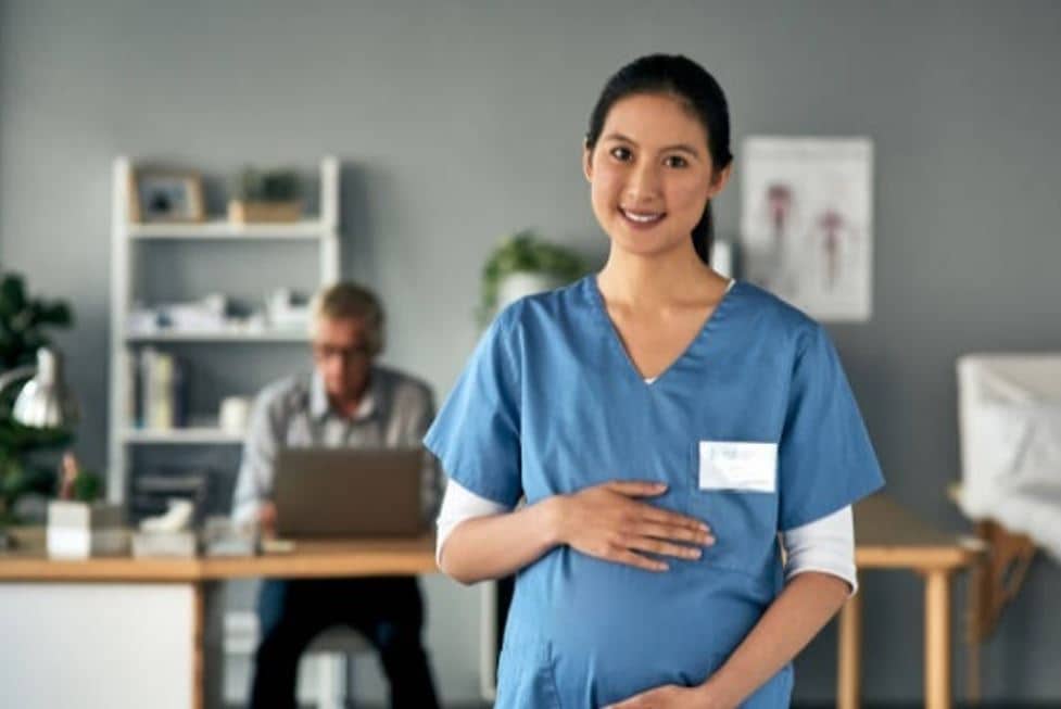 Pregnant woman wearing scrubs and holding her belly with a doctor in a white coat sits in the background at the computer