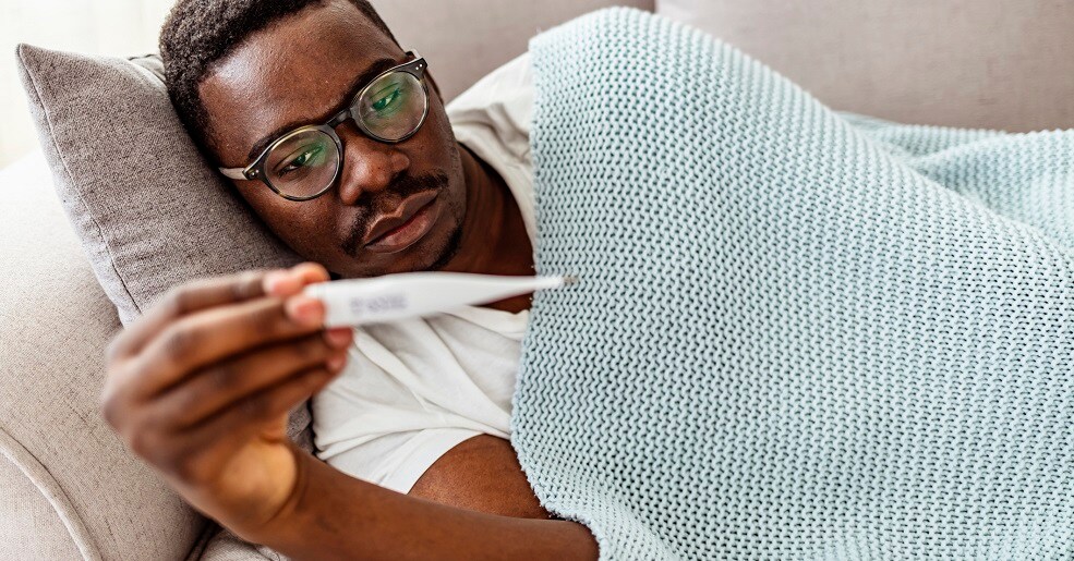 Man laying on couch, sick, and taking temperature.