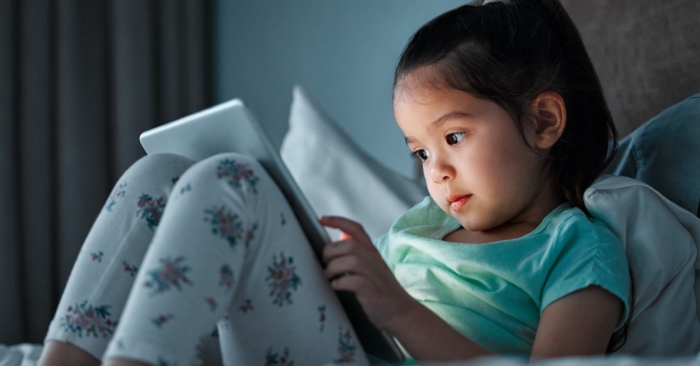 Girl sitting in dark on top of a blanket, looking at tablet.