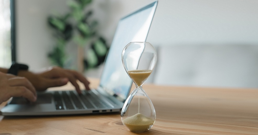 Laptop in use at home office, next to a hour glass on desk.