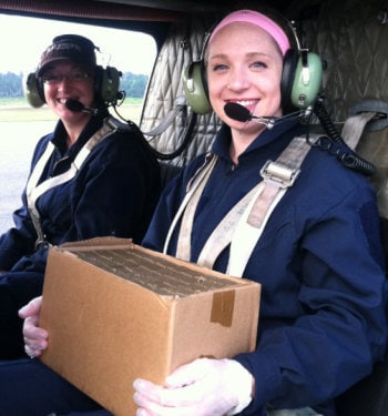 Two young women in a helicopter, one of them wearing gloves and carrying a sealed box of supplies.