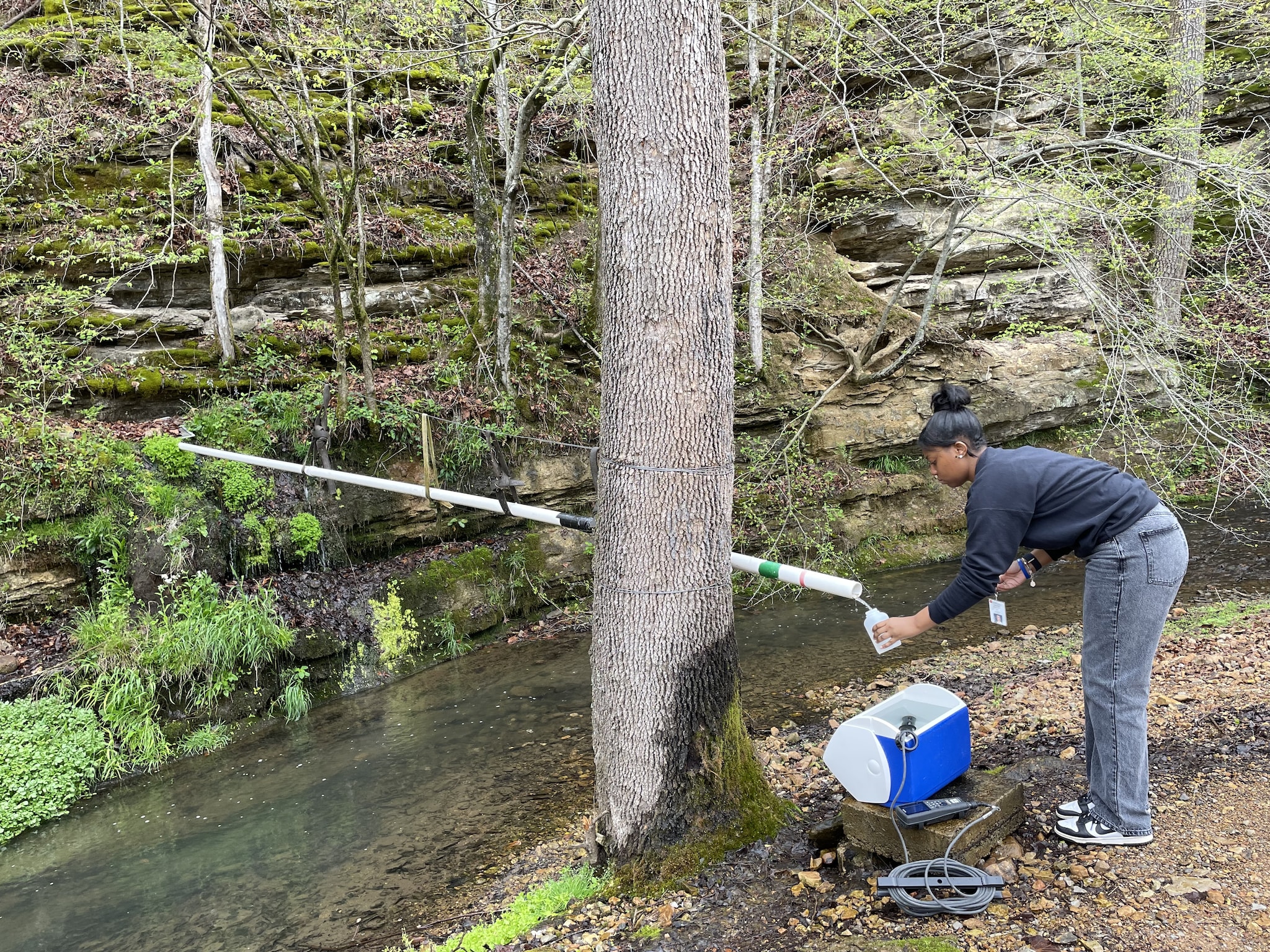 A person stands next to a flowing creek. They hold a bottle collecting water from a pipe on the opposite bank.