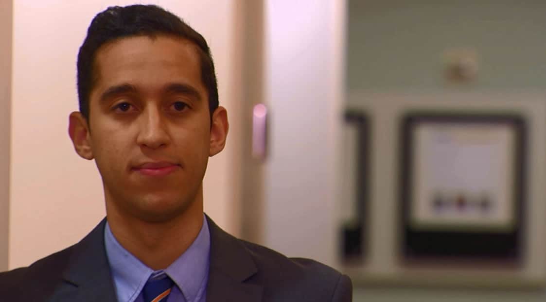 A young man in a suit and tie confidently gazes at the camera in a blurred office setting, exuding professionalism and poise.