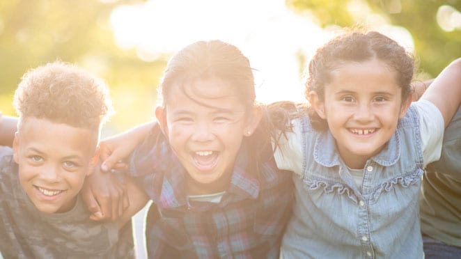 Three young children outdoors, arms around each other, and smiling.