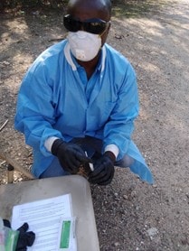 person in a lab coat, mask, glove and protective eye gear squats down on the ground near some paperwork