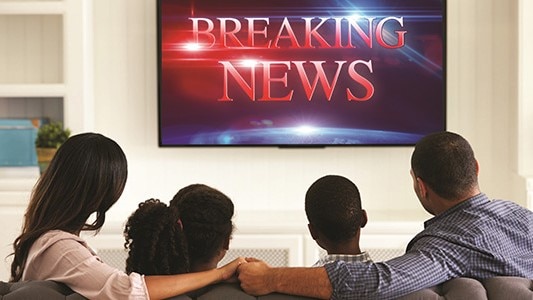 A family on their living room sofa watching the TV. The TV screen says “Breaking News.