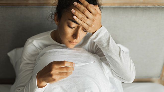 Una mujer enferma sentada en la cama mirando un termómetro.