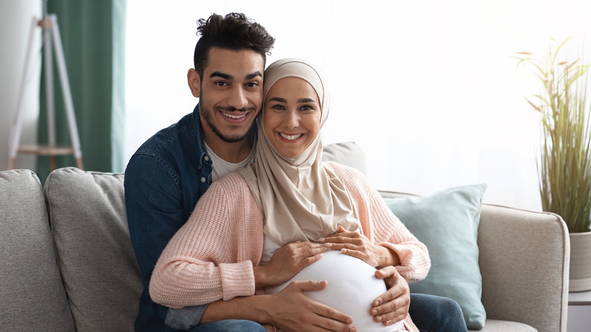 Pregnant woman wearing hijab sitting on couch with man embracing her from behind with hands on her belly.