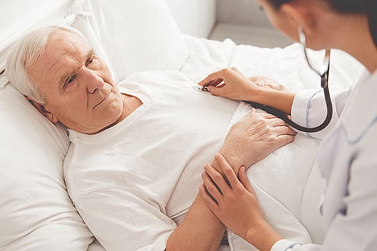 Older adult patient lies in bed as doctor presses stethoscope onto chest