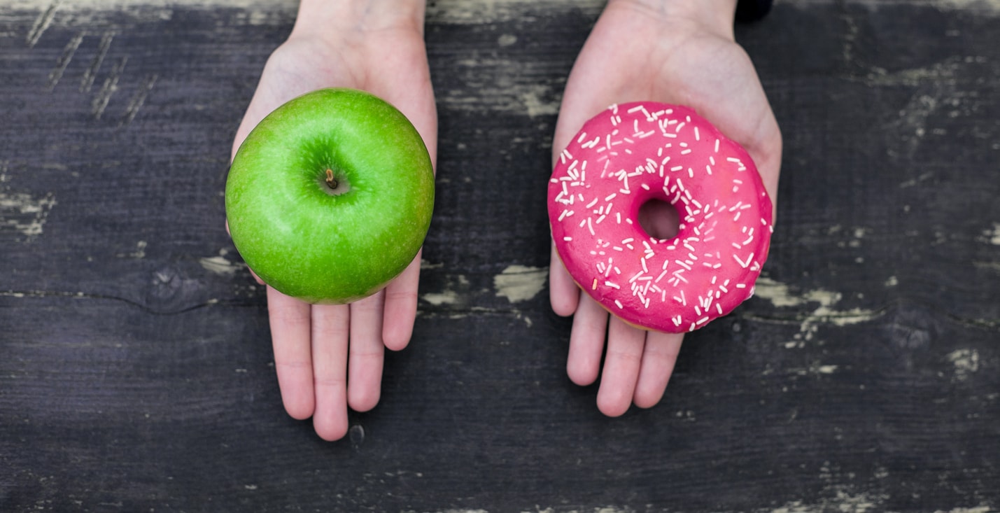 Two open hands. One hand is holding a green apple. The other hand is holding a pink donut with sprinkles.