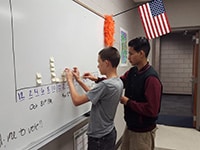 Two diverse students engage in a lesson on a whiteboard in a classroom setting.