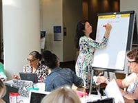 A dark-haired woman in a colorful dress writes on a giant notepad in a room filled with diverse peers.