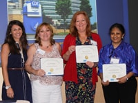 A diverse group of four women standing in front of a projected graphic holding certificates.