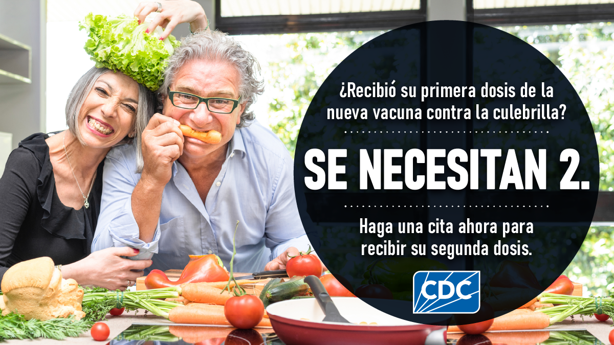 Una pareja sonriendo en la cocina con frutas y verduras en la mesa. El texto dice: El texto dice: ¿Recibió su primera dosis de la nueva vacuna contra la culebrilla? Se necesitan 2. Haga una cita ahora para recibir su segunda dosis.