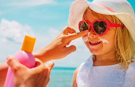 A una niña con sombrero de ala ancha y gafas de sol se le aplica protector solar en la cara. A una niña con sombrero de ala ancha y gafas de sol se le aplica protector solar en la cara.
