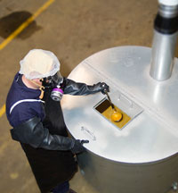 2012-148.jpg_d9379 Recipiente para mezclar con controles de ingeniería (tapa de un tanque con bisagras y ventilación, con tapa de acceso). Trabajador con el equipo de protección personal apropiado puesto (respirador que cubre la cara completa con purificador de aire equipado con cartuchos para vapor orgánico P100, delantal y guantes).