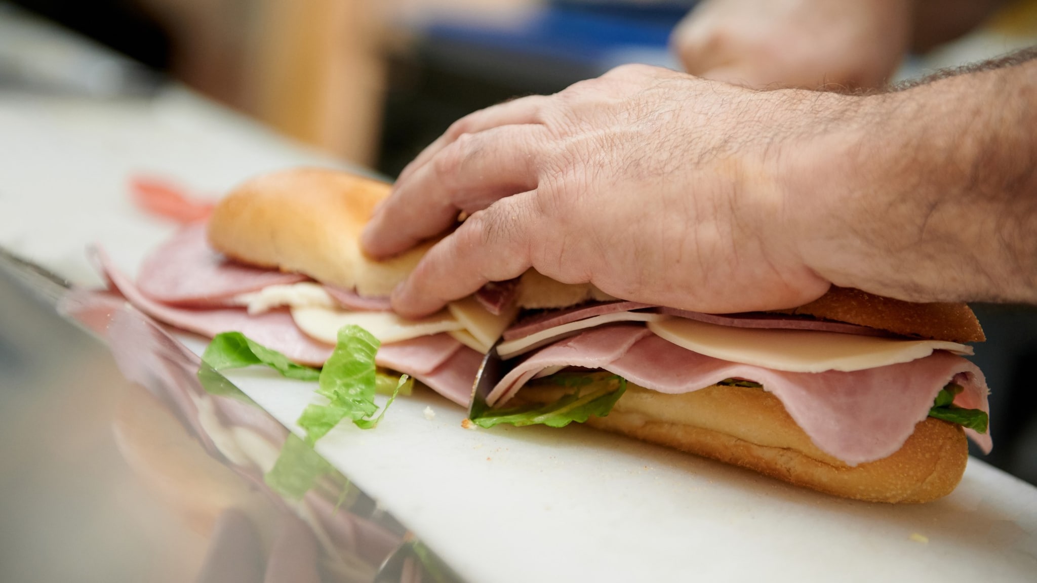 A person preparing a deli meat sub sandwich on a white cutting board.
