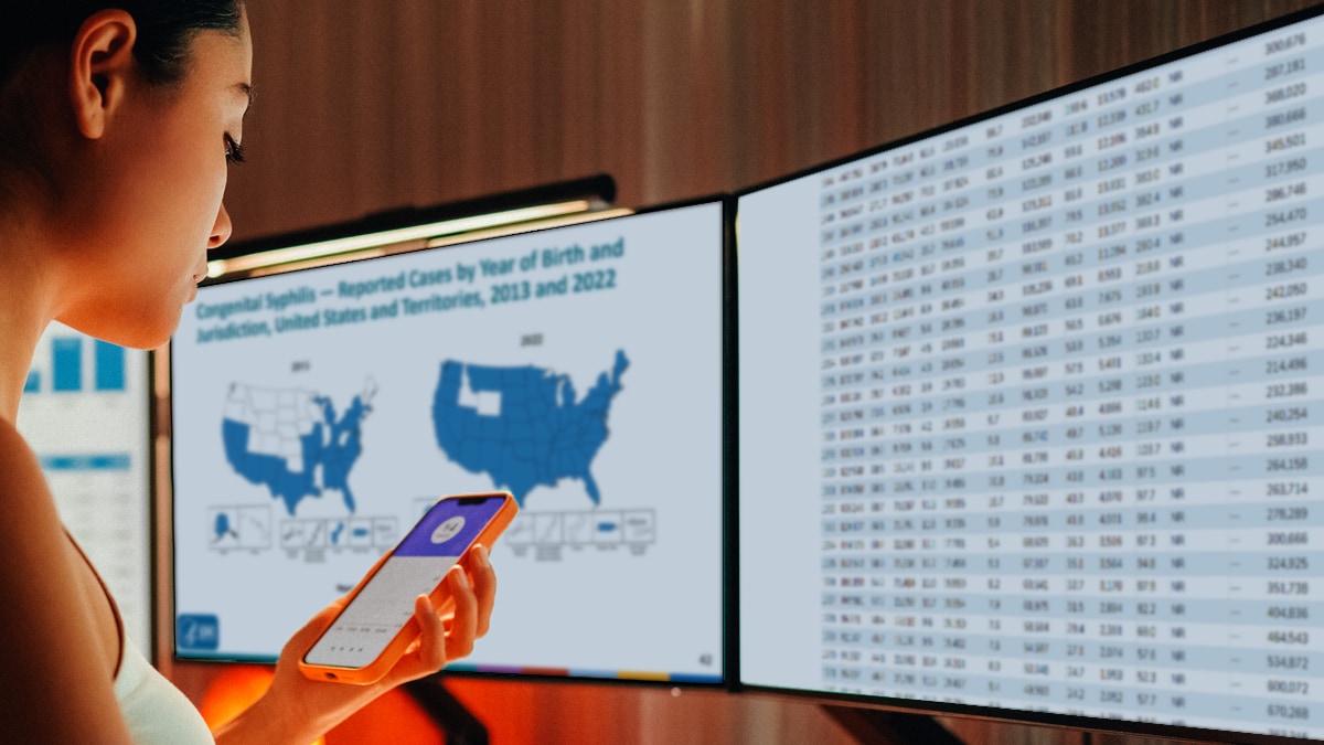 A woman with a handheld device and two monitors showing statistical content.