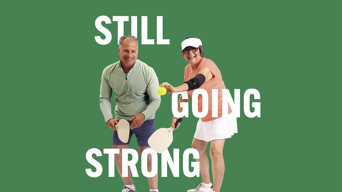 An older man and woman, wearing athletic clothing, are holding pickleball paddles and the woman is about to serve the ball.