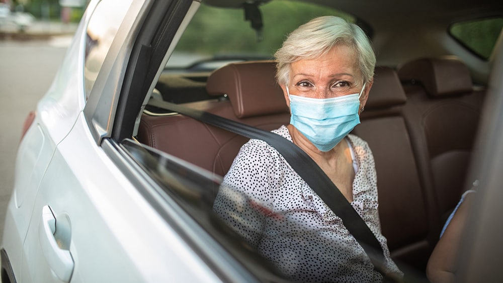 Older woman sitting in the back seat of a car and wearing a protective face mask