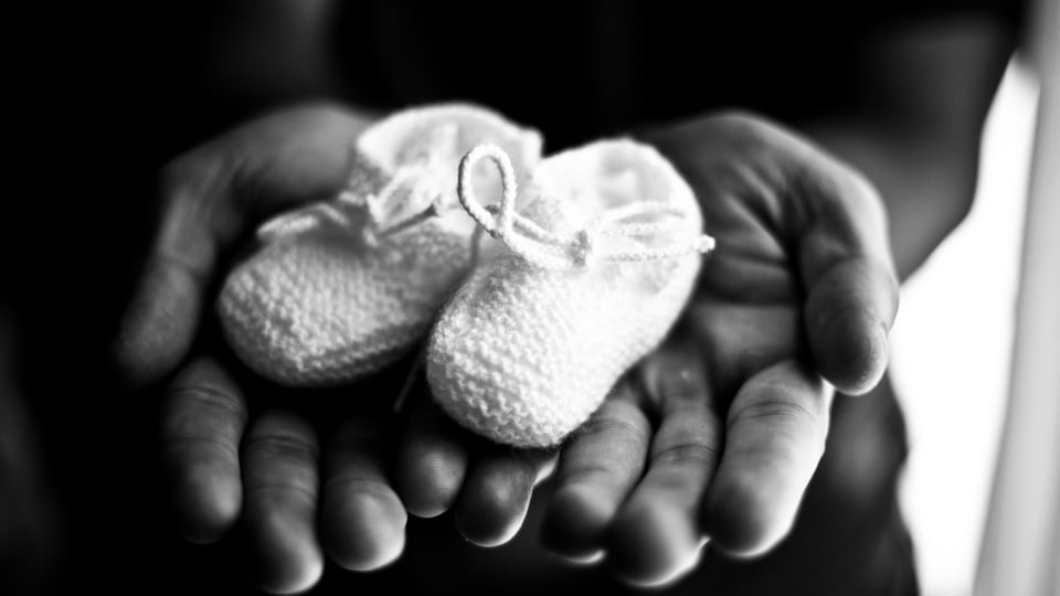 Foto en blanco y negro de las manos de un adulto sosteniendo un par de zapatos de bebé tejidos.