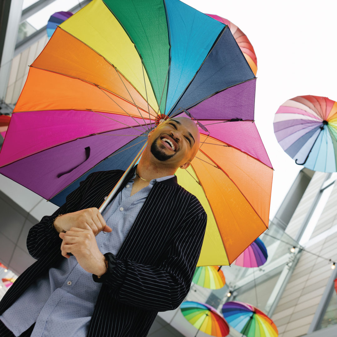 Pride – Jay An image of a man smiling outdoors with rainbow umbrellas in the background.