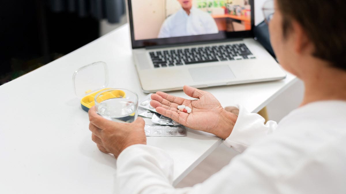 Woman holding pills and a glass of water and talking to a health care provider on a laptop.