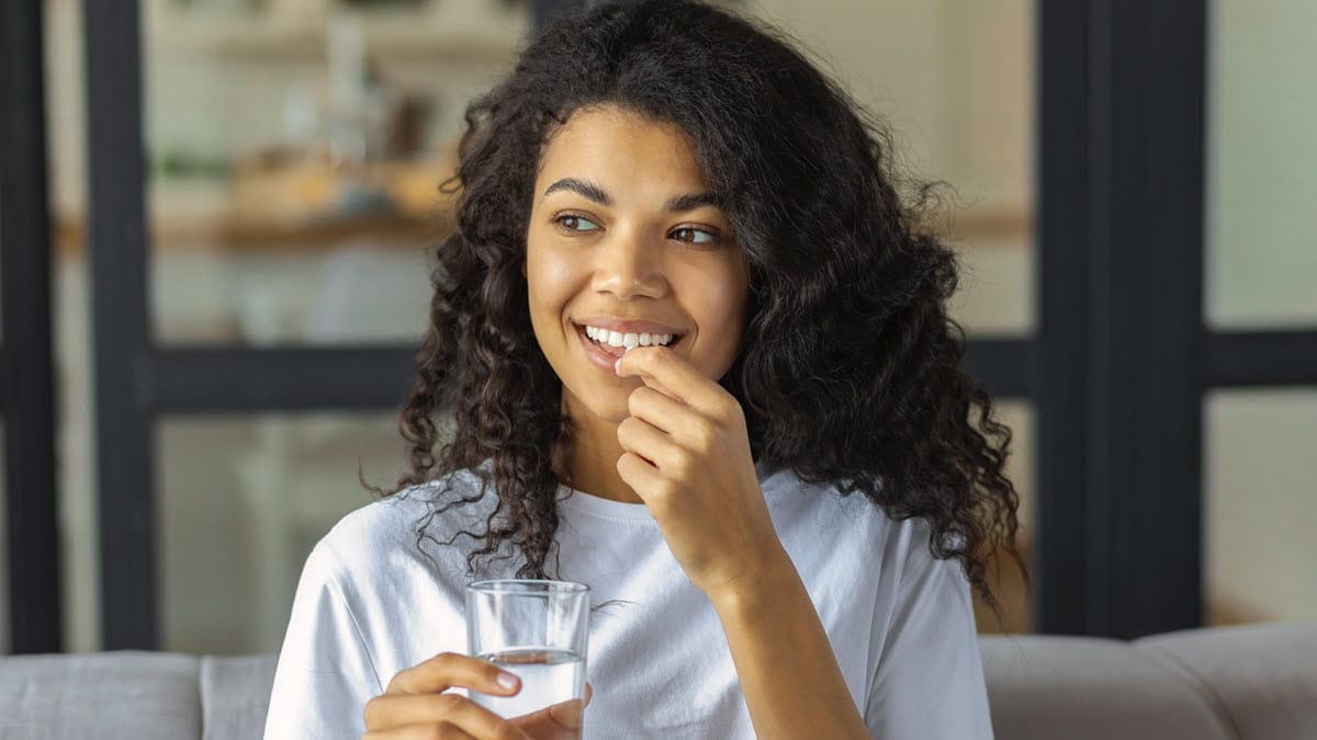 Young African American woman smiling and taking medicine while holding a glass of water.