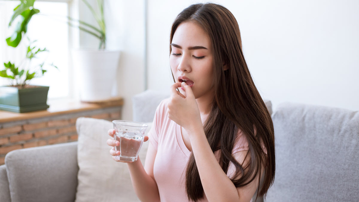 A woman takes pills with a glass of water while sitting on the sofa