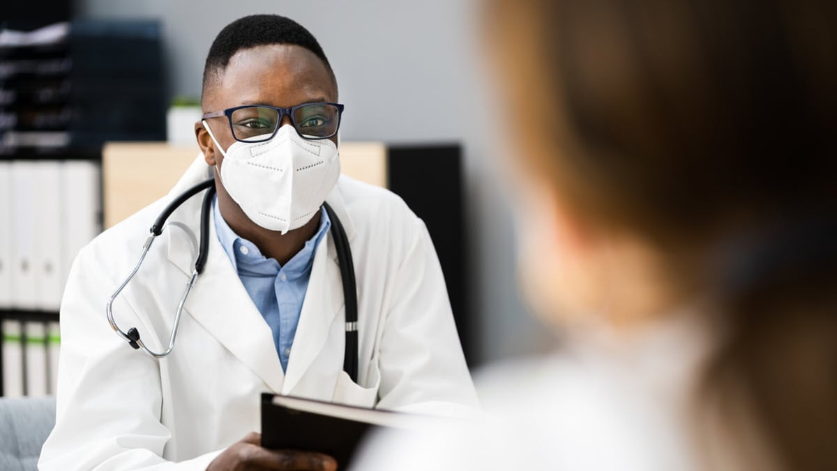 A male health care provider wearing a respiratory speaks to a female patient.