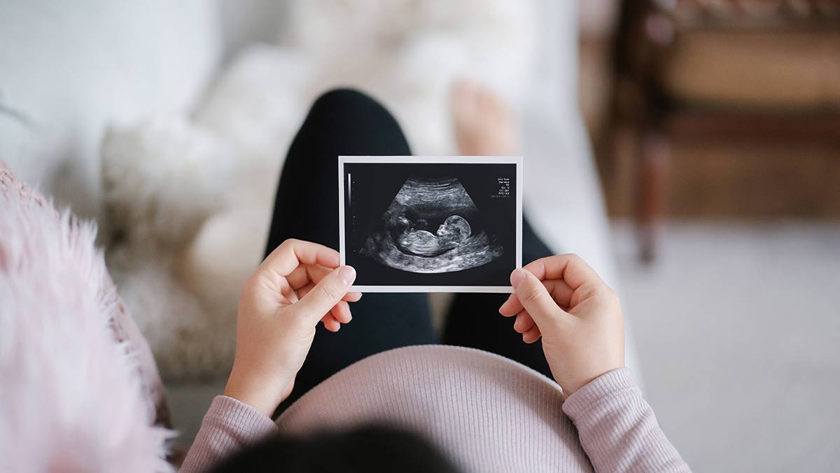 Young asian pregnant woman lying on sofa at home looking at the ultrasound scan photo of her baby