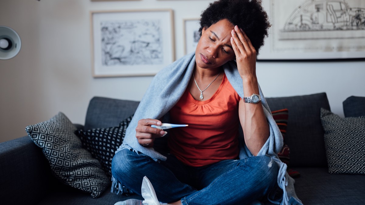 A woman feeling ill while sitting on a couch views an oral thermometer.