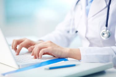 A close-up of a healthcare professional’s hands typing on a laptop keyboard. The person is wearing a white coat with a stethoscope around their neck.