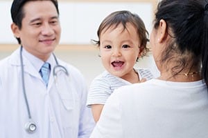 A woman with dark hair is holding a smiling baby as she talks with a dark haired male healthcare provider