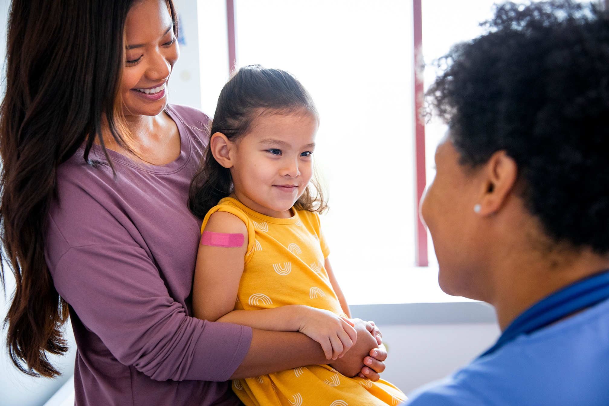 Pediatric patient with band-aid on upper arm sitting with parent talking to healthcare provider in a medical office.