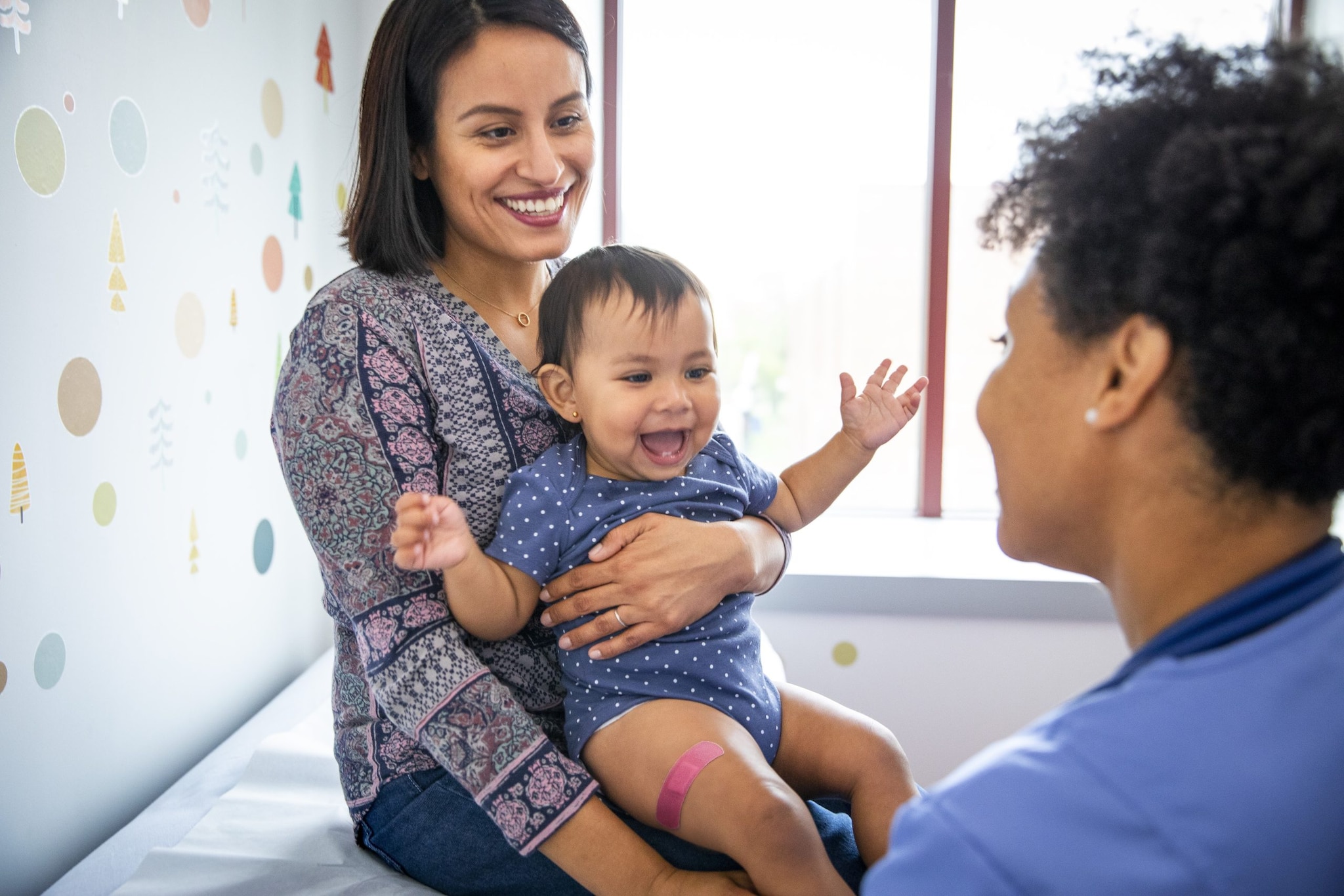 Parent holding infant with a band aid on leg in medical office with healthcare provider post vaccination.