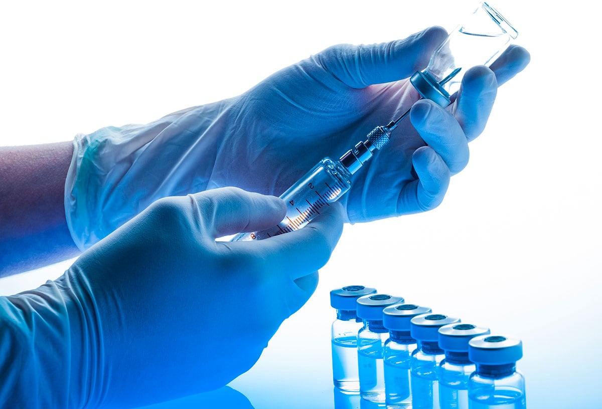 Preparing vaccine: Hands with gloves holding syringe and vial. Blue toned image. Selective focus. Several vials are out of focus on background on foreground and two vials are out of focus at background.