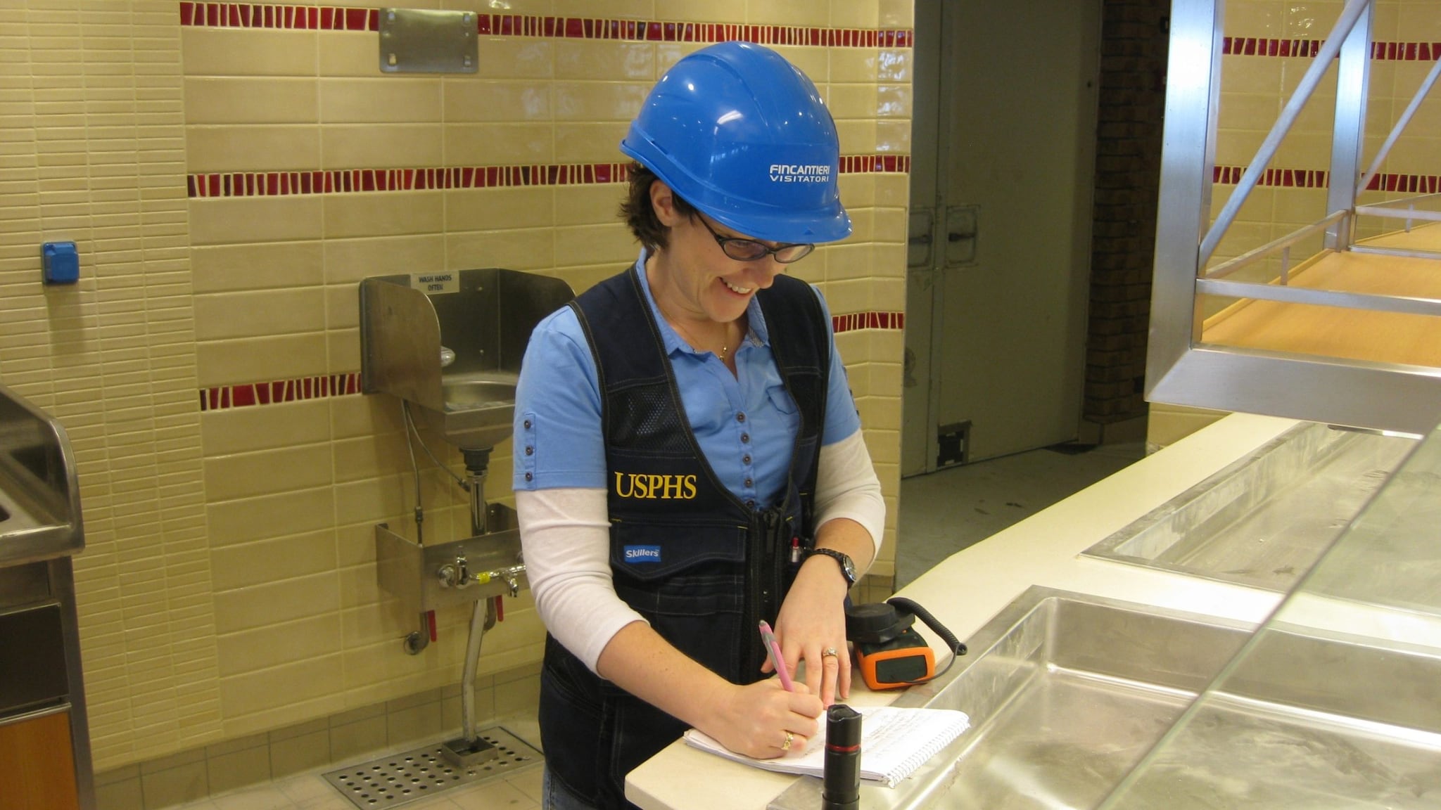 Public health officer wearing a hard hat taking notes on a clipboard inside a galley.