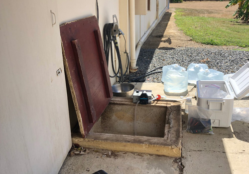 Una cisterna subterránea externa al lado de una edificación. La tapa de madera está abierta y se ve la abertura de concreto cuadrada. Hay una nevera portátil, bolsas de agua y un instrumento para pruebas del agua alrededor de la abertura de la cisterna.