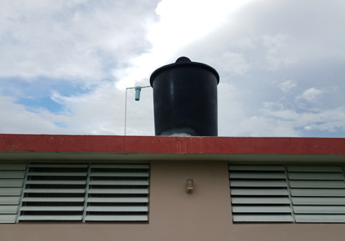 Edificación con un recipiente de almacenamiento de agua grande (cisterna) en el techo. Hay un tubo de plástico entre la cisterna y el techo, con un filtro de agua conectado al tubo.