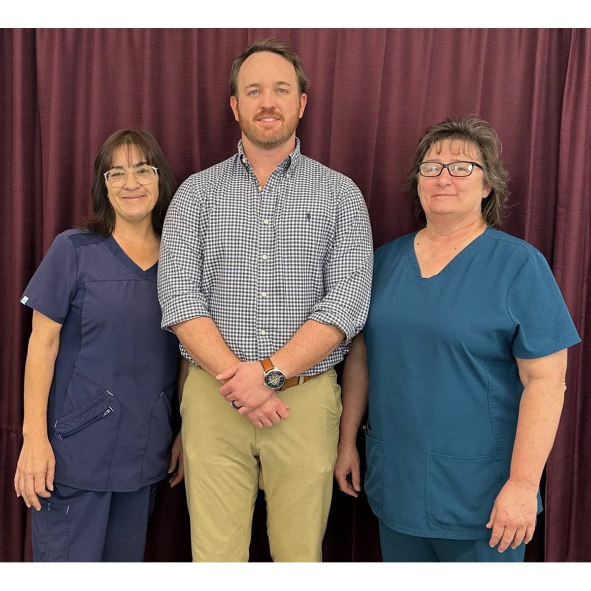 2 women wearing scrubs and a tall man from Faith Healthcare smiling in front of a red curtain